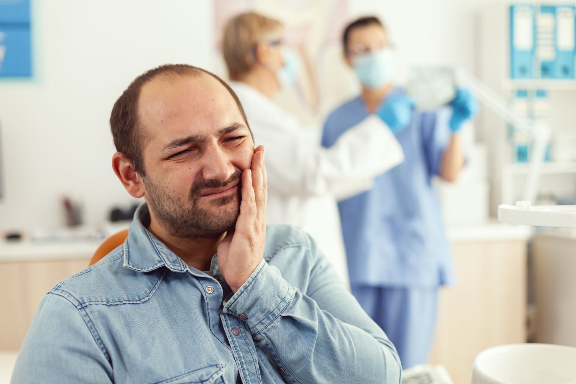 Man holding his cheek in a dental clinic, grimacing while staff in the background talk