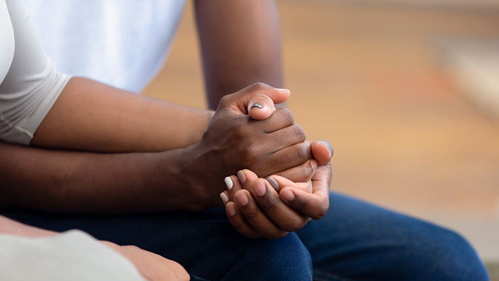 Hands clasped together, one person's darker skin cradled by another's lighter skin; comfort and support.