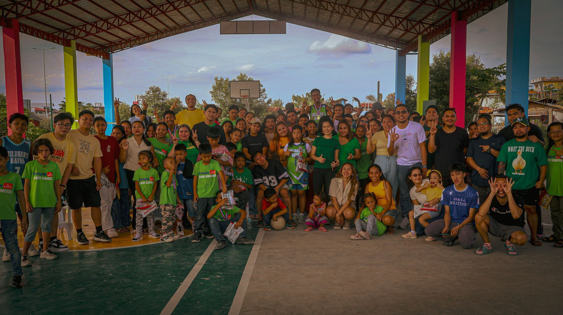 A large group of people, many in green shirts, pose on a basketball court under a covered area, smiling and gesturing.