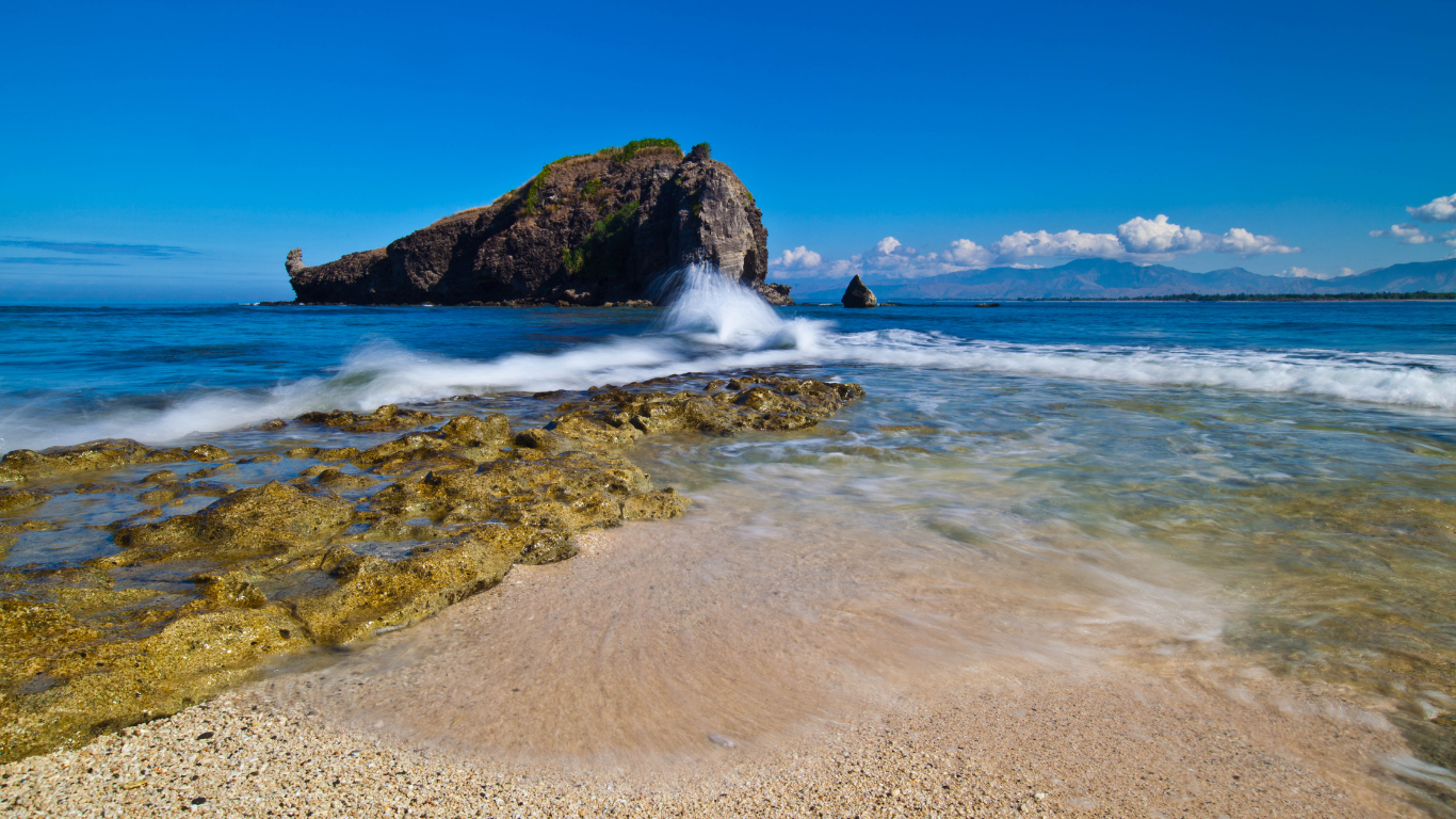 Sandy beach with waves crashing over rocks, leading to a rocky island under a clear blue sky.