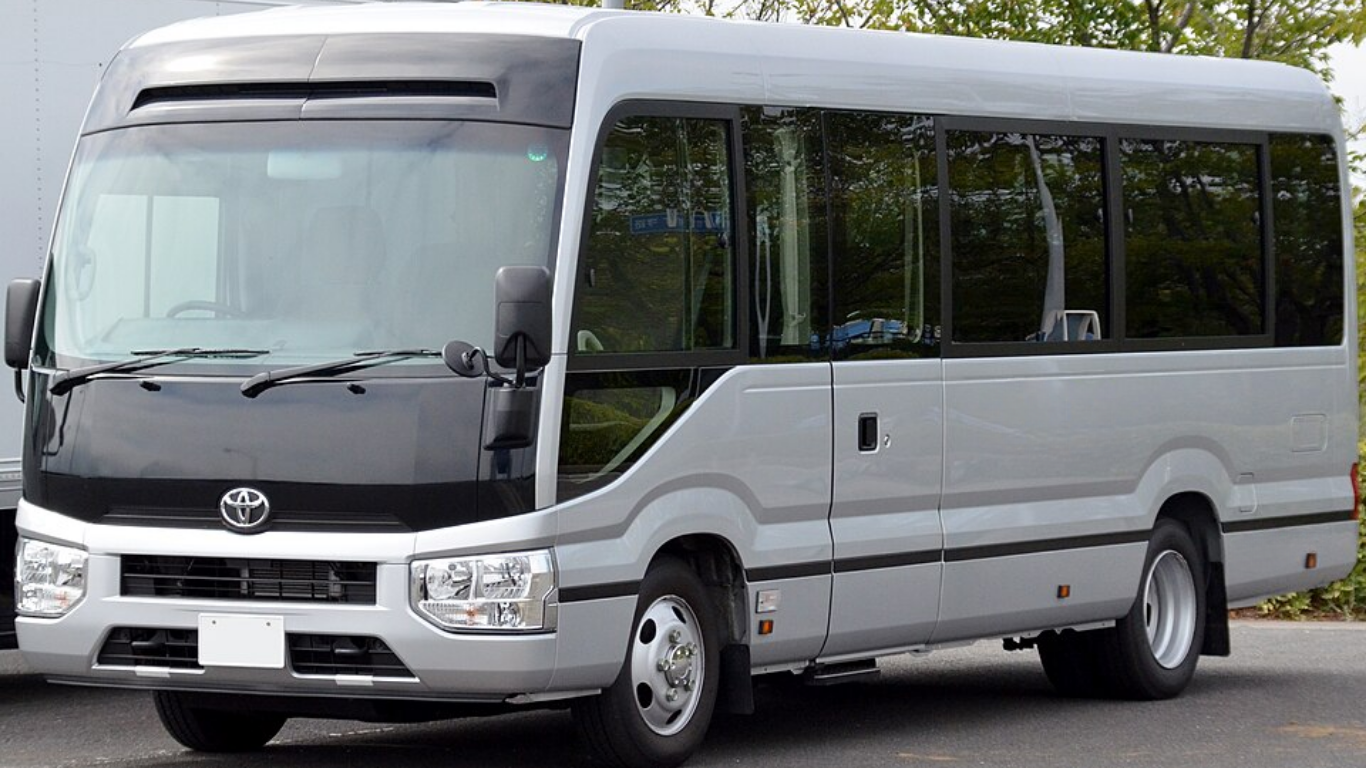 Silver Toyota Coaster minibus with black roof, parked outside on a paved area.