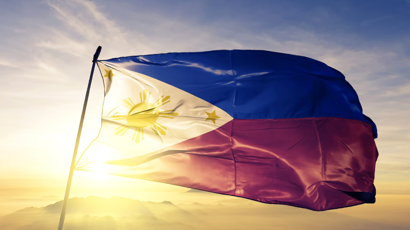 Philippine flag waving in the wind against a sunny, golden sky; features blue and red stripes, a white triangle with a sun and stars.