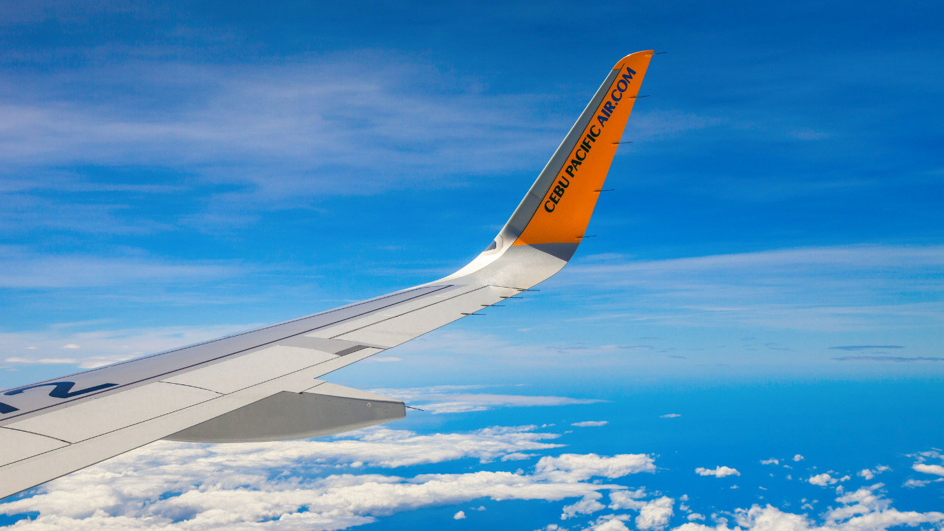 Airplane wing against a bright blue sky with fluffy white clouds. The wing's tip has orange and black accents.