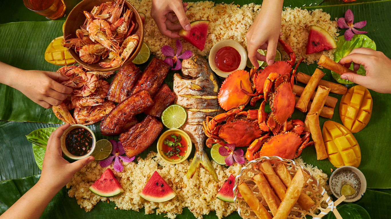 People's hands reaching for a bountiful feast on banana leaves: seafood, pork, rice, fruit, and sauces are spread out for sharing.