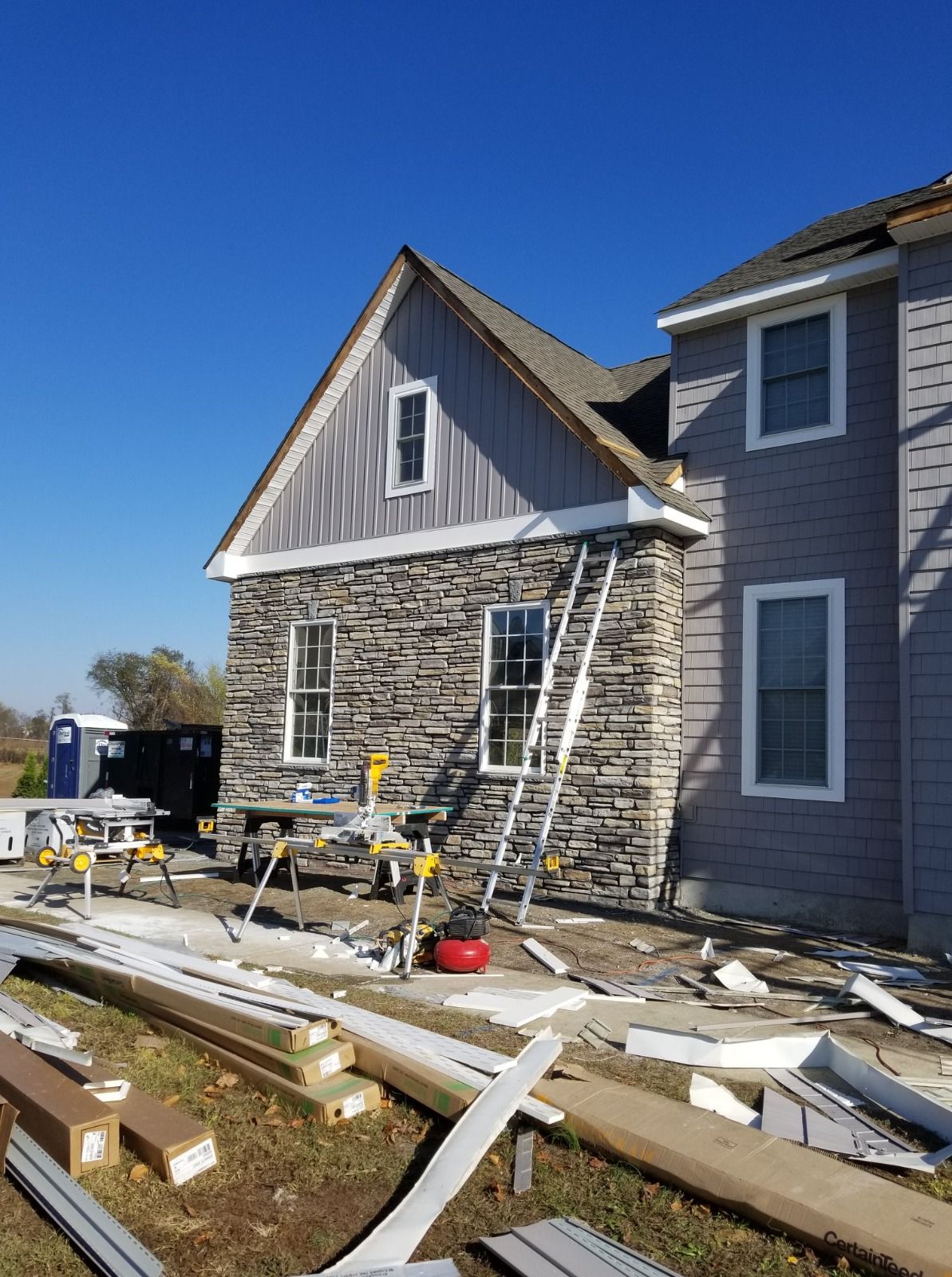 Construction of a house exterior with stone and siding. A ladder leans against the stone wall. Blue sky.