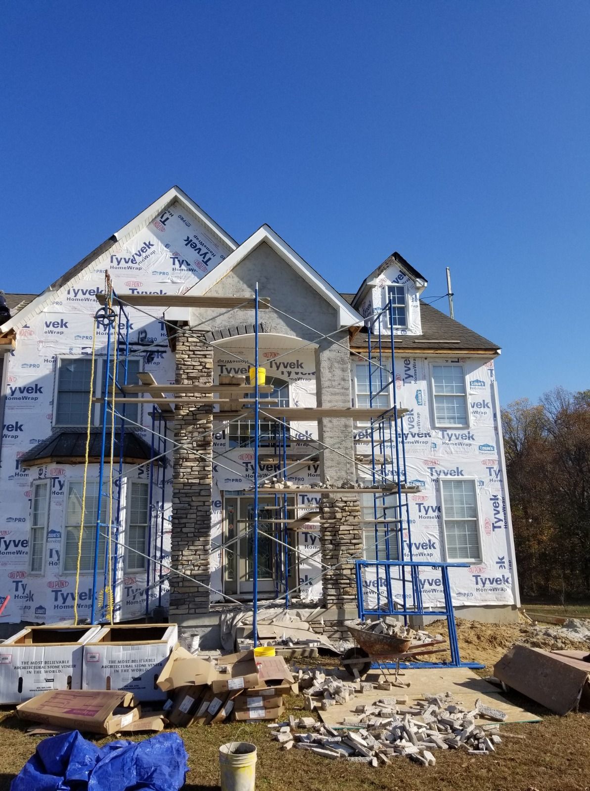 House under construction, with scaffolding, blue wrap, and stonework being applied to the exterior under a clear sky.