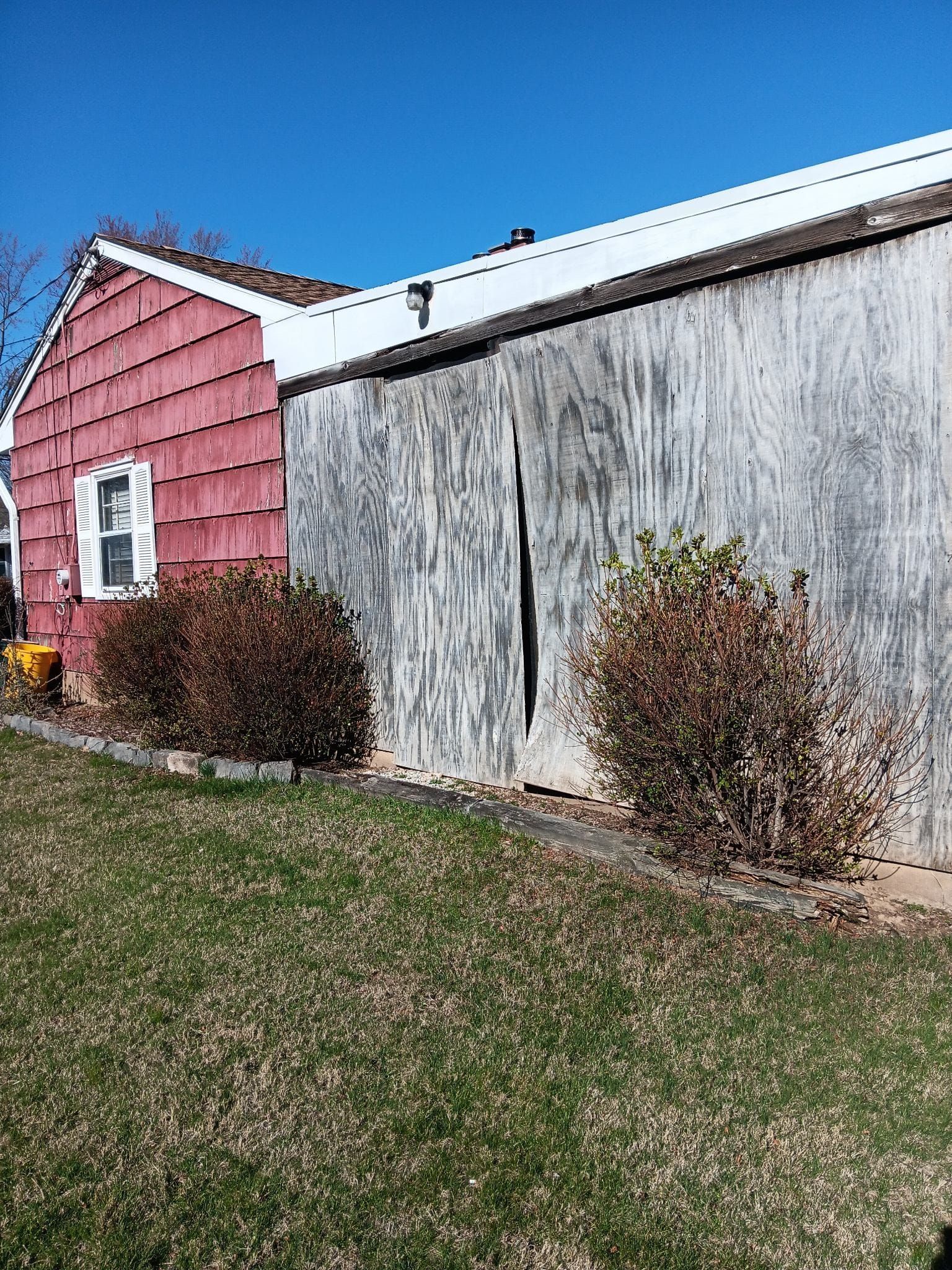 Red house with white-trimmed window next to a weathered, gray barn, on a grassy hill under a blue sky.