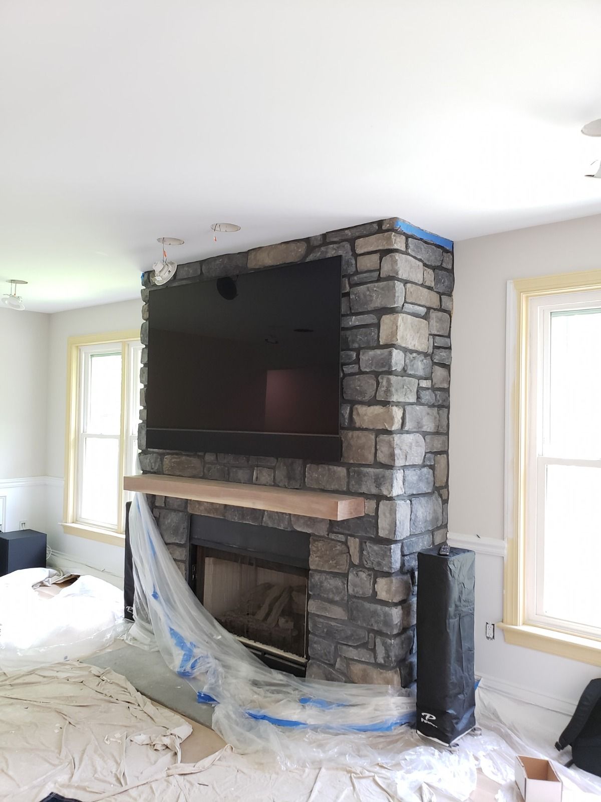 Stone fireplace with mounted TV, mantle, and windows. Blue painter's tape and plastic sheeting present.