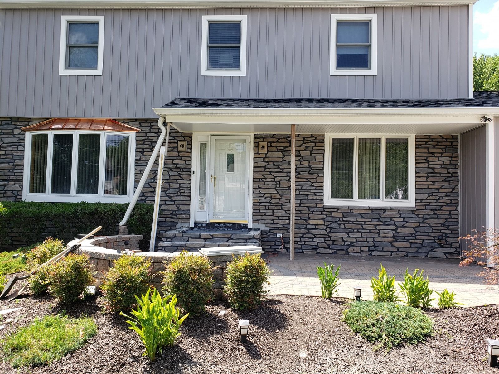 Two-story house with gray siding, stone facade, and white trim. Front entrance with steps, bushes, and windows.