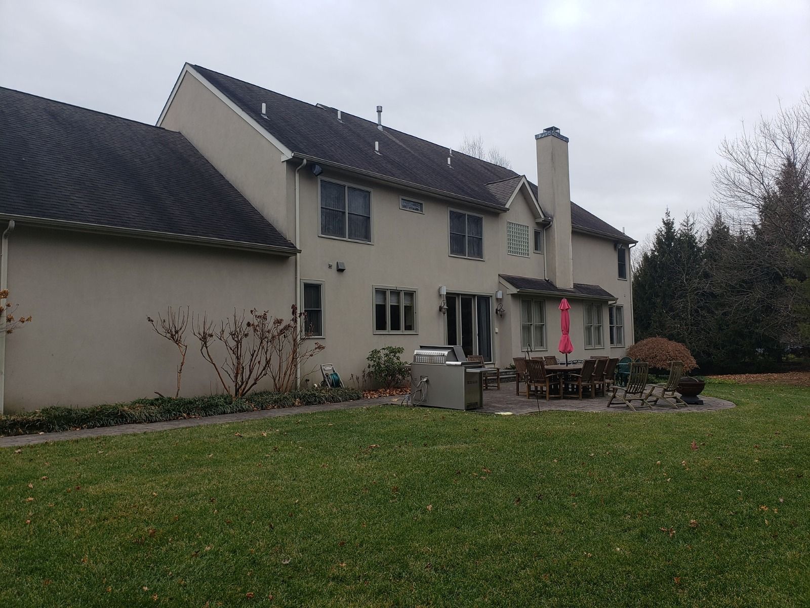 Beige two-story house with dark roof and chimney, backyard with patio, lawn, and trees under overcast sky.