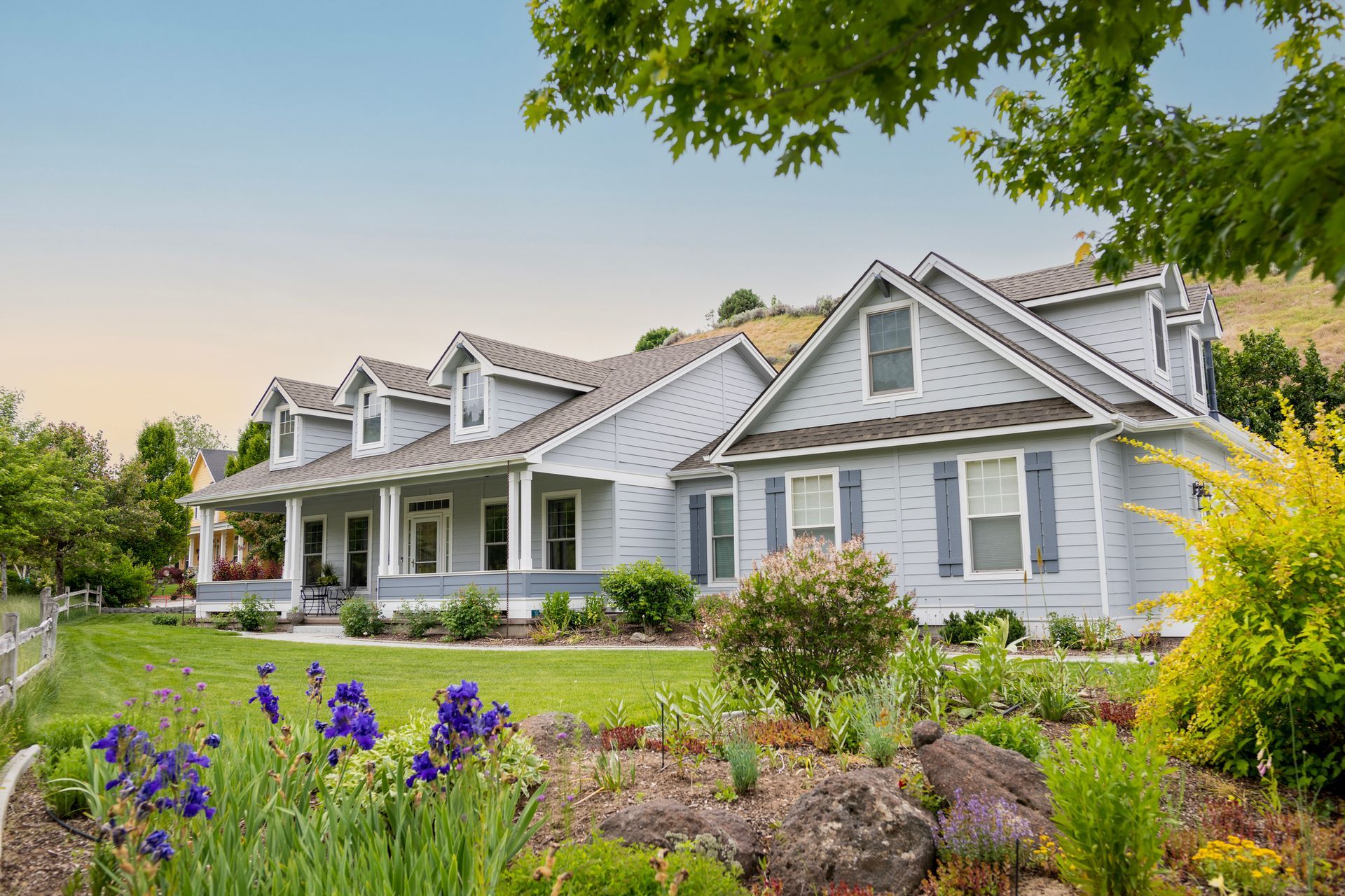 Blue house with porch and dormers, garden with purple flowers, green grass, and trees.