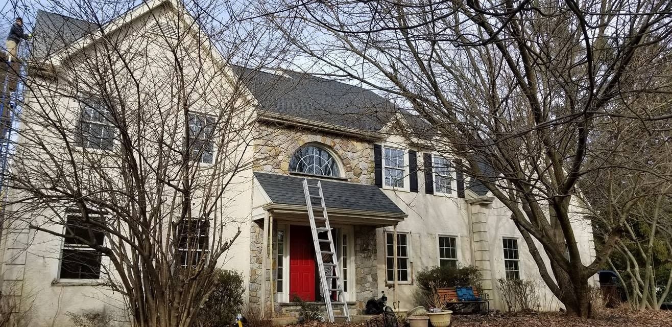 House with a red front door, gray roof, and stone facade; ladder propped against the roof. Trees in foreground.