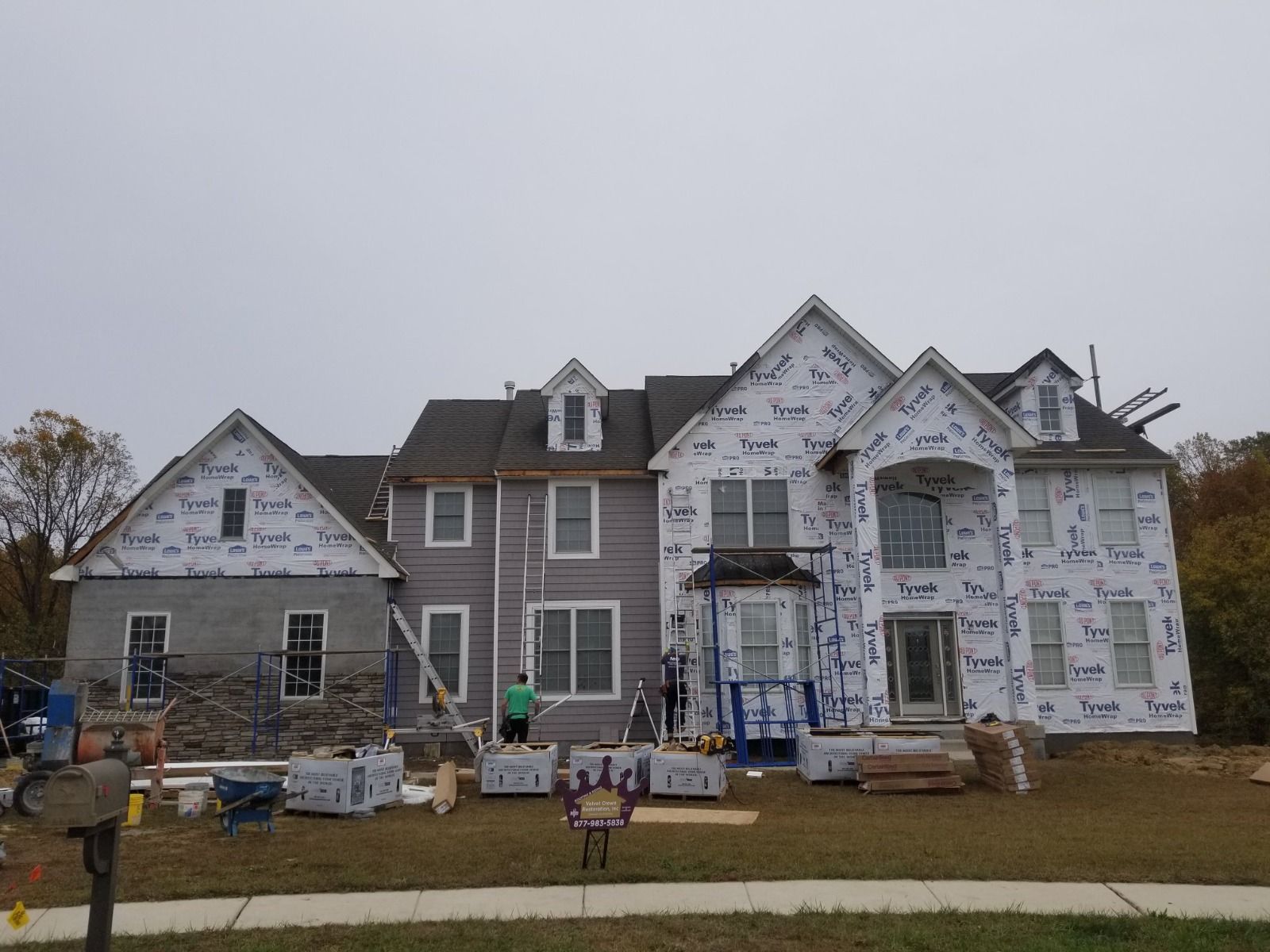 House under construction, covered in wrap. Blue scaffolding, workers, and materials on the lawn
