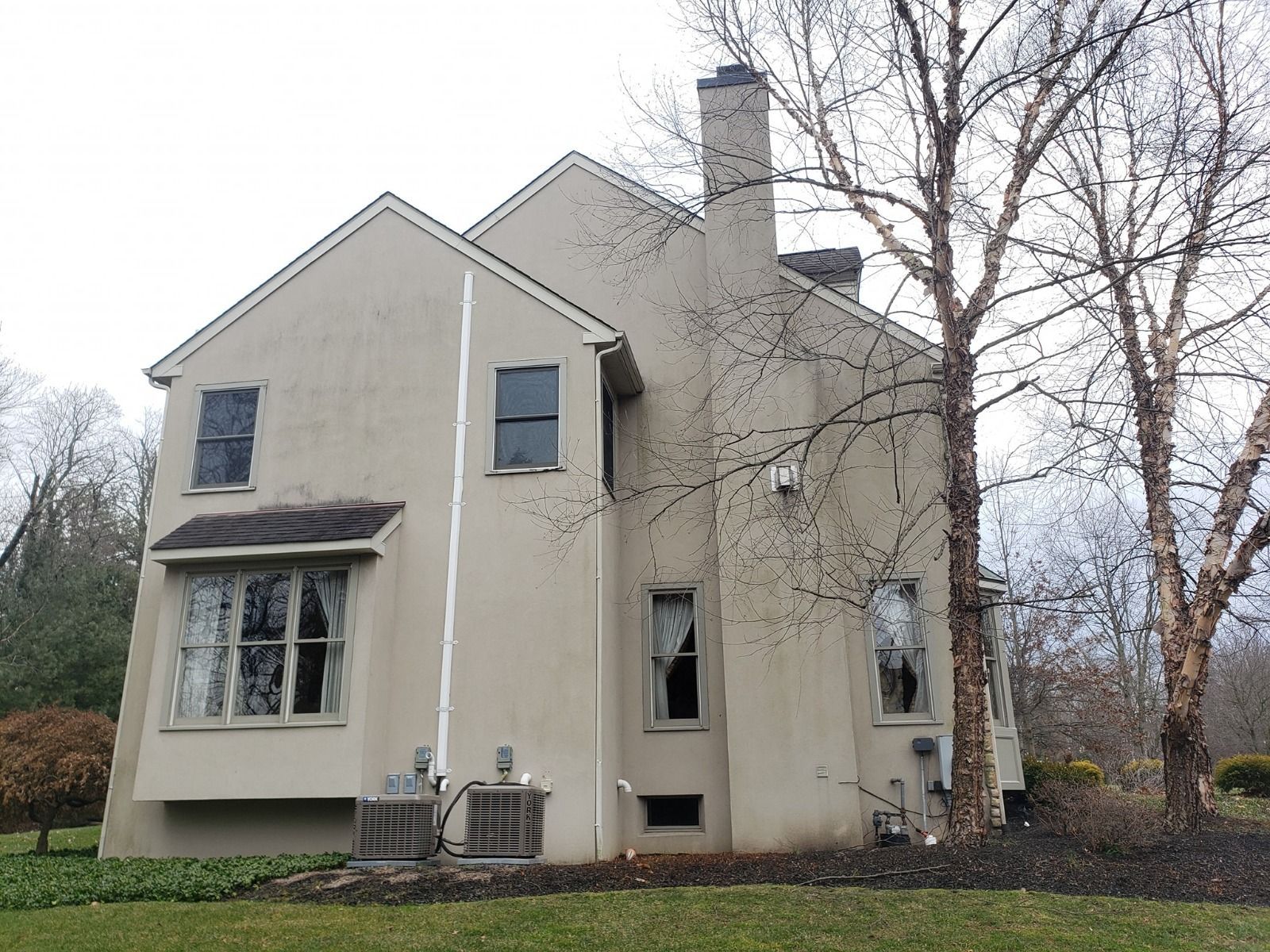 Beige two-story house with a chimney and several windows, set on a green lawn with bare trees.