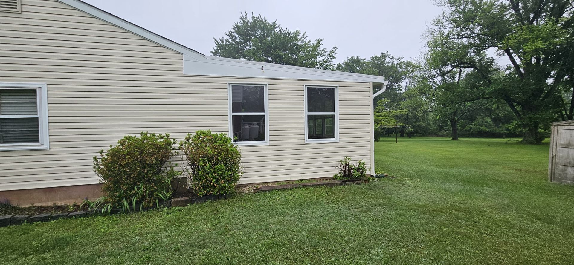 A house with light siding and two windows, surrounded by green grass and trees.