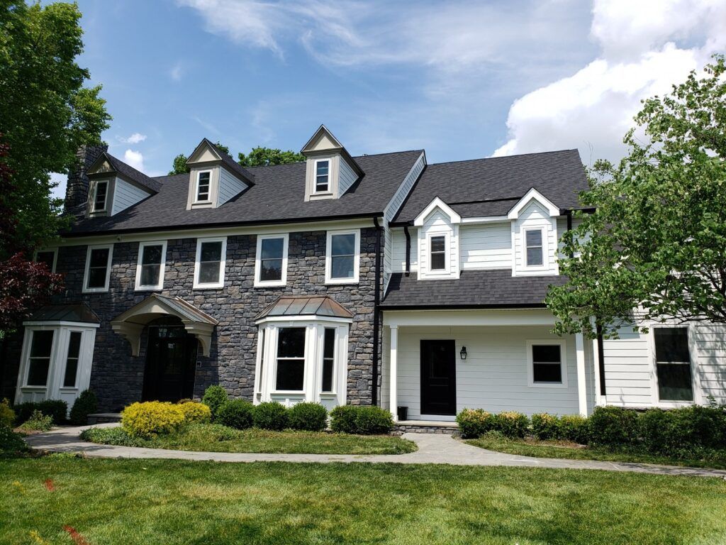 Two-story house with stone and white siding, black roof, and dormer windows, on a green lawn.