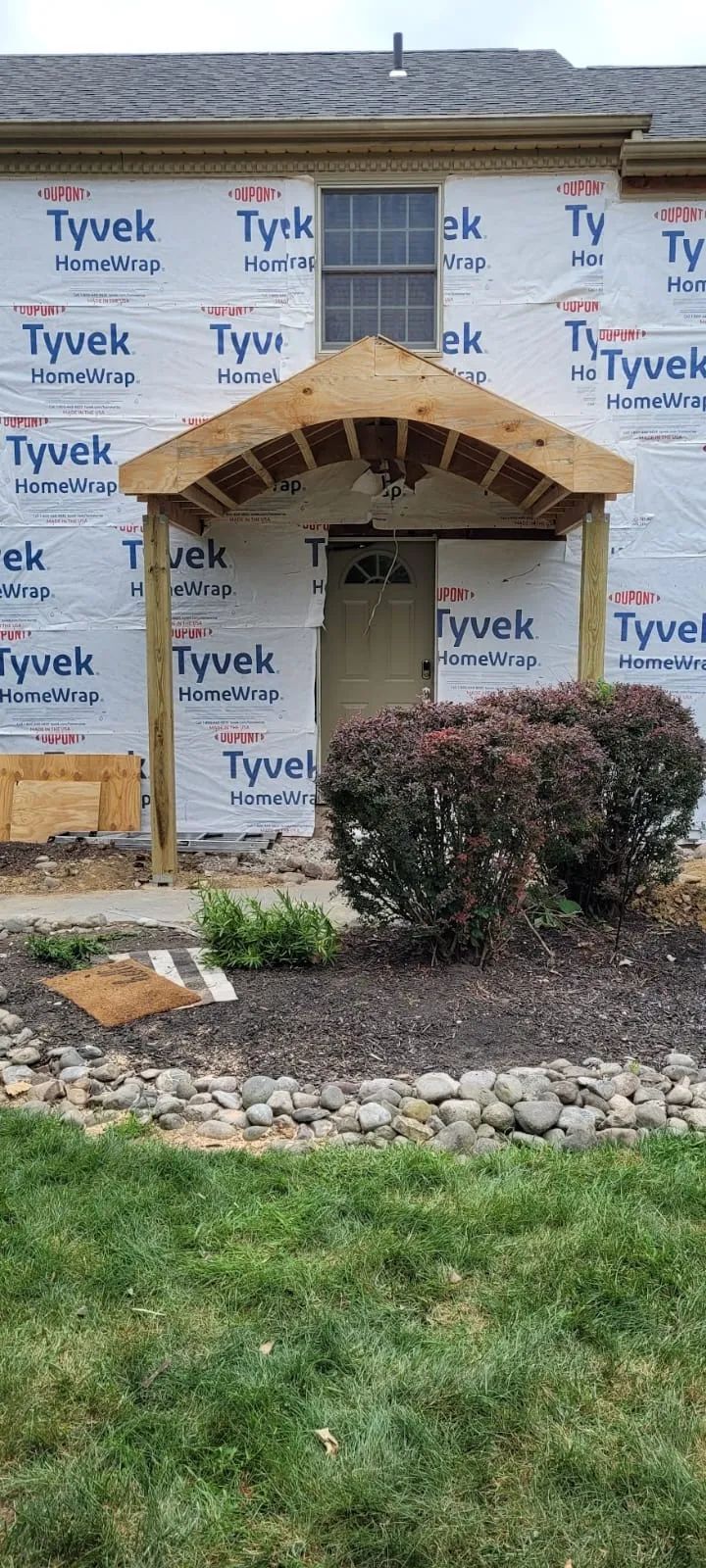 House under construction; Tyvek wrap, wooden entryway under a small roof, overgrown bushes in front.