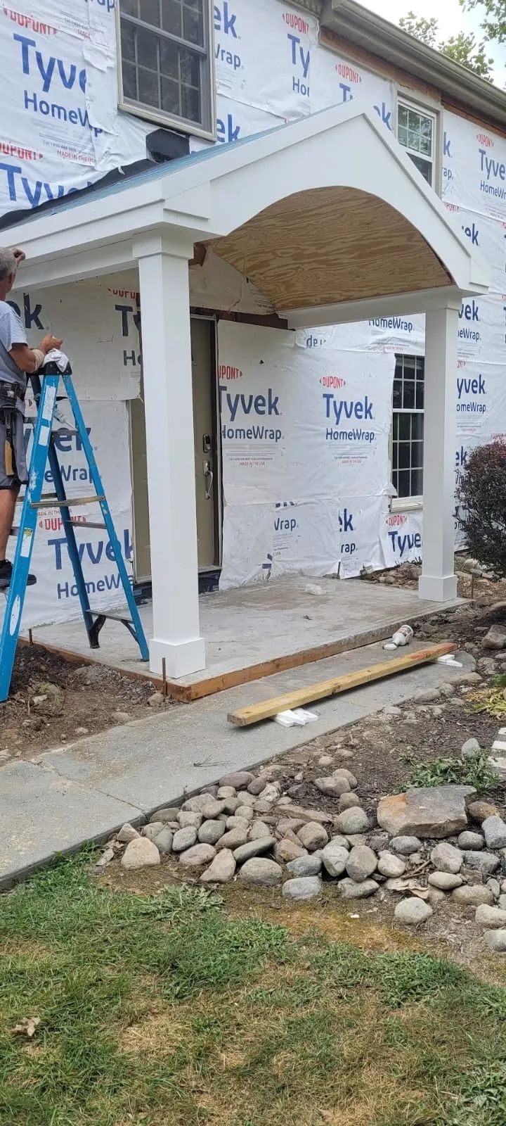 Construction of a porch with white columns. A worker on a ladder. House exterior is covered with Tyvek wrap.