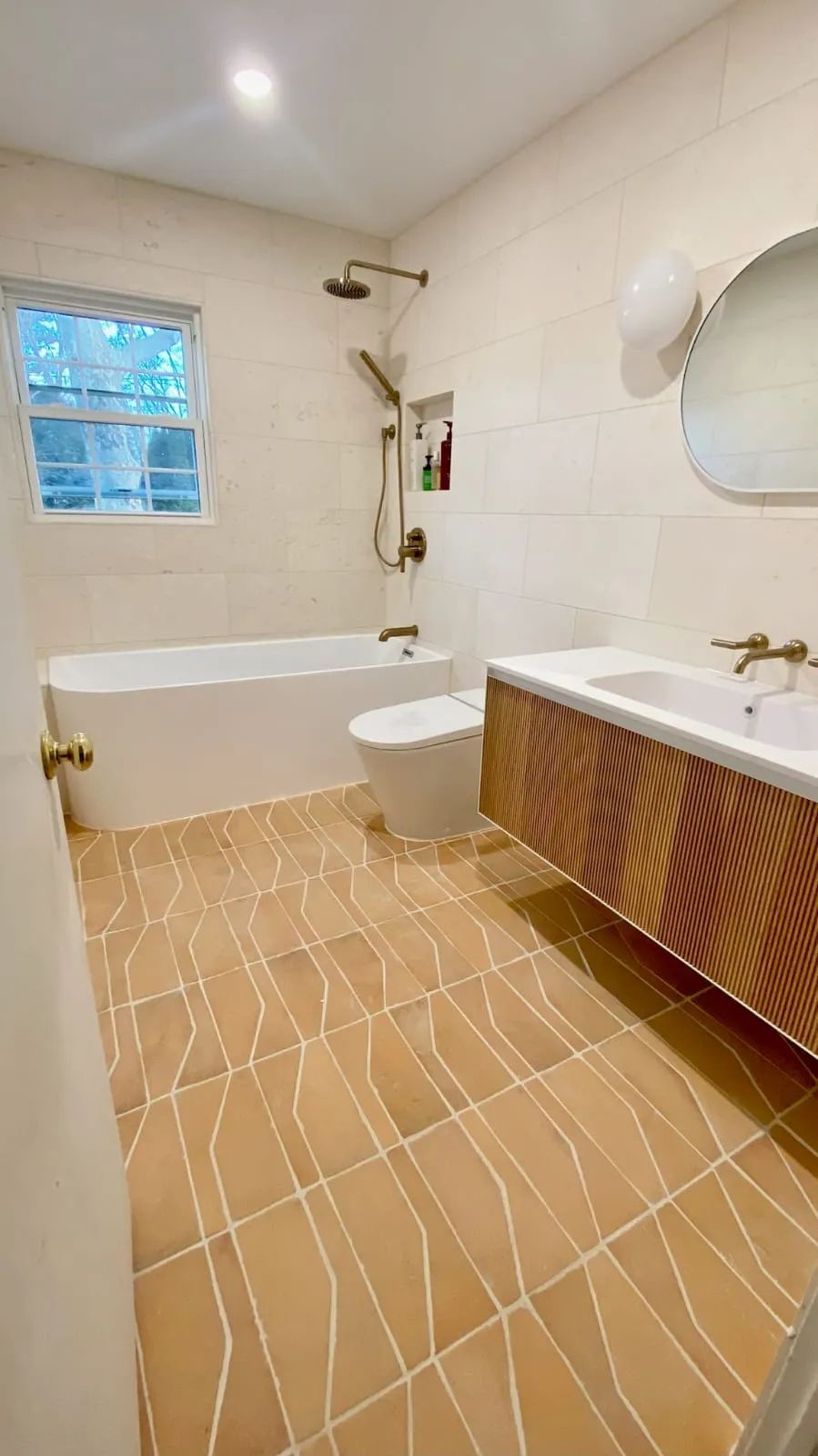 Modern bathroom with white tub, toilet, and sink, and wood vanity. Tan tile floor and wall.