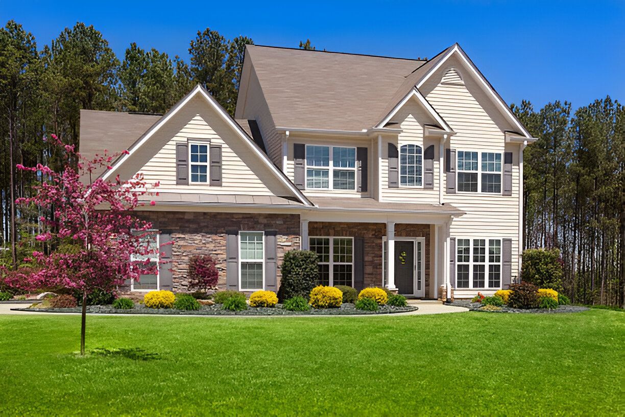 Two-story house with beige siding, stone accents, and brown roof, set on green lawn with blue sky and trees.