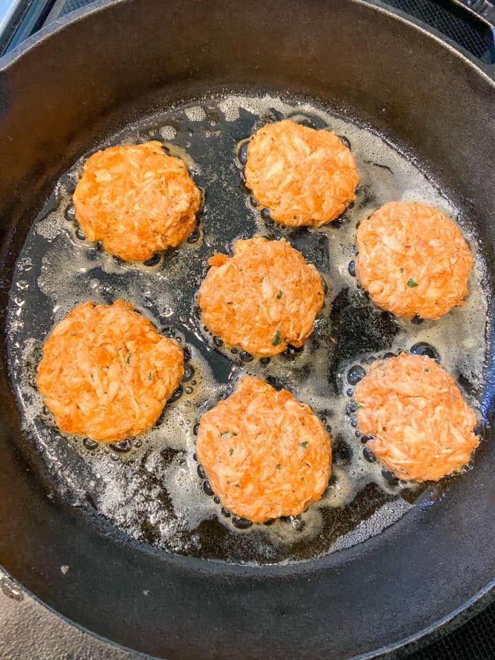 Six golden-brown, circular fritters frying in a cast iron skillet with bubbling oil.