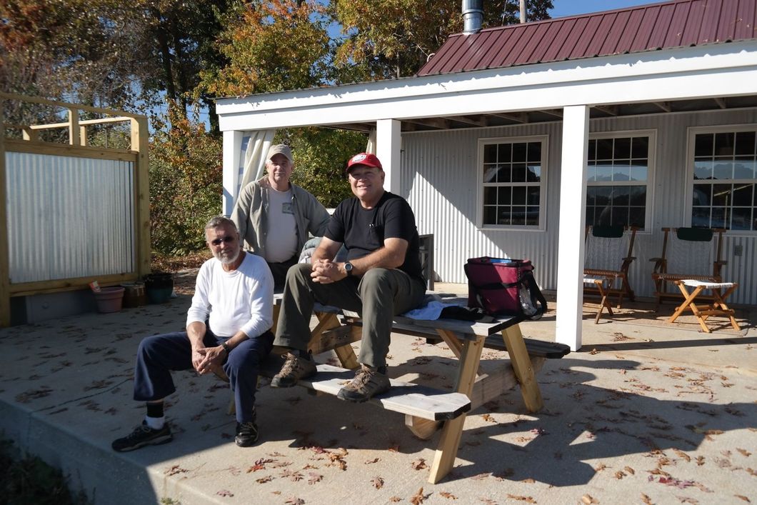 Three people sit at a picnic table on a porch outside a white building with a maroon roof on a sunny autumn day.