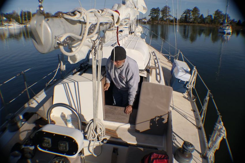 A person wearing a beanie and grey sweatshirt stands in the cockpit of a sailboat on calm, sunny water.