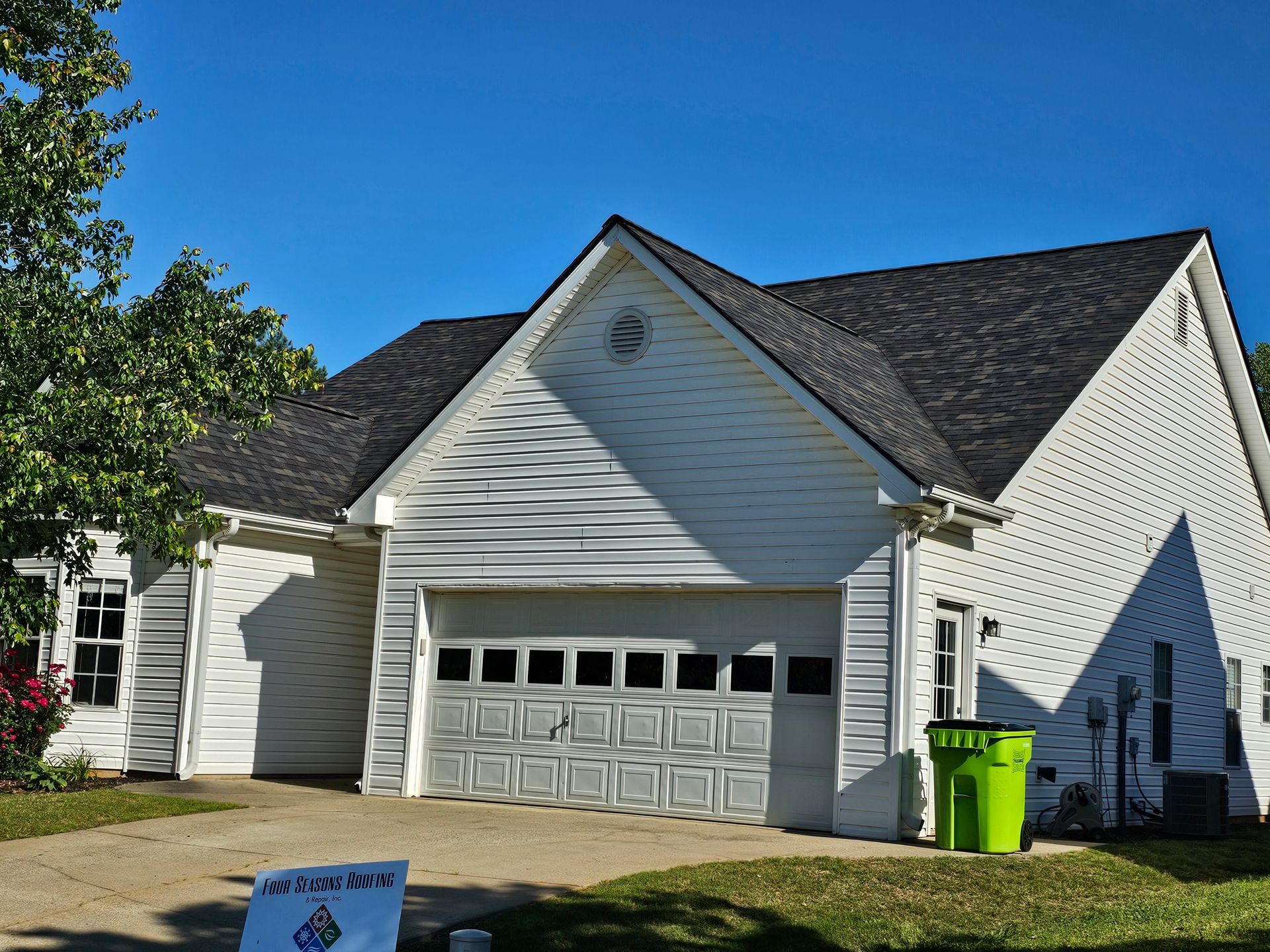 White house with a garage, green trash can, and new black roof under a blue sky.