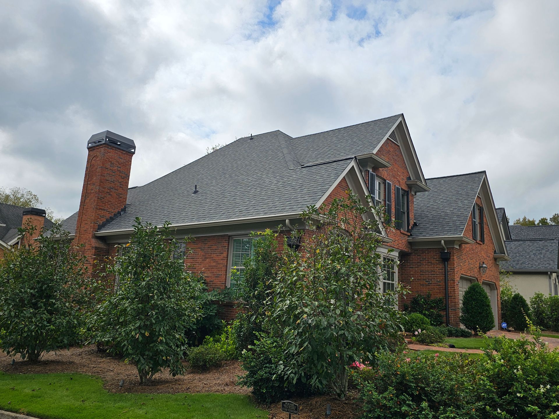 Brick house with a dark gray roof, chimney, and bushes. Overcast sky.