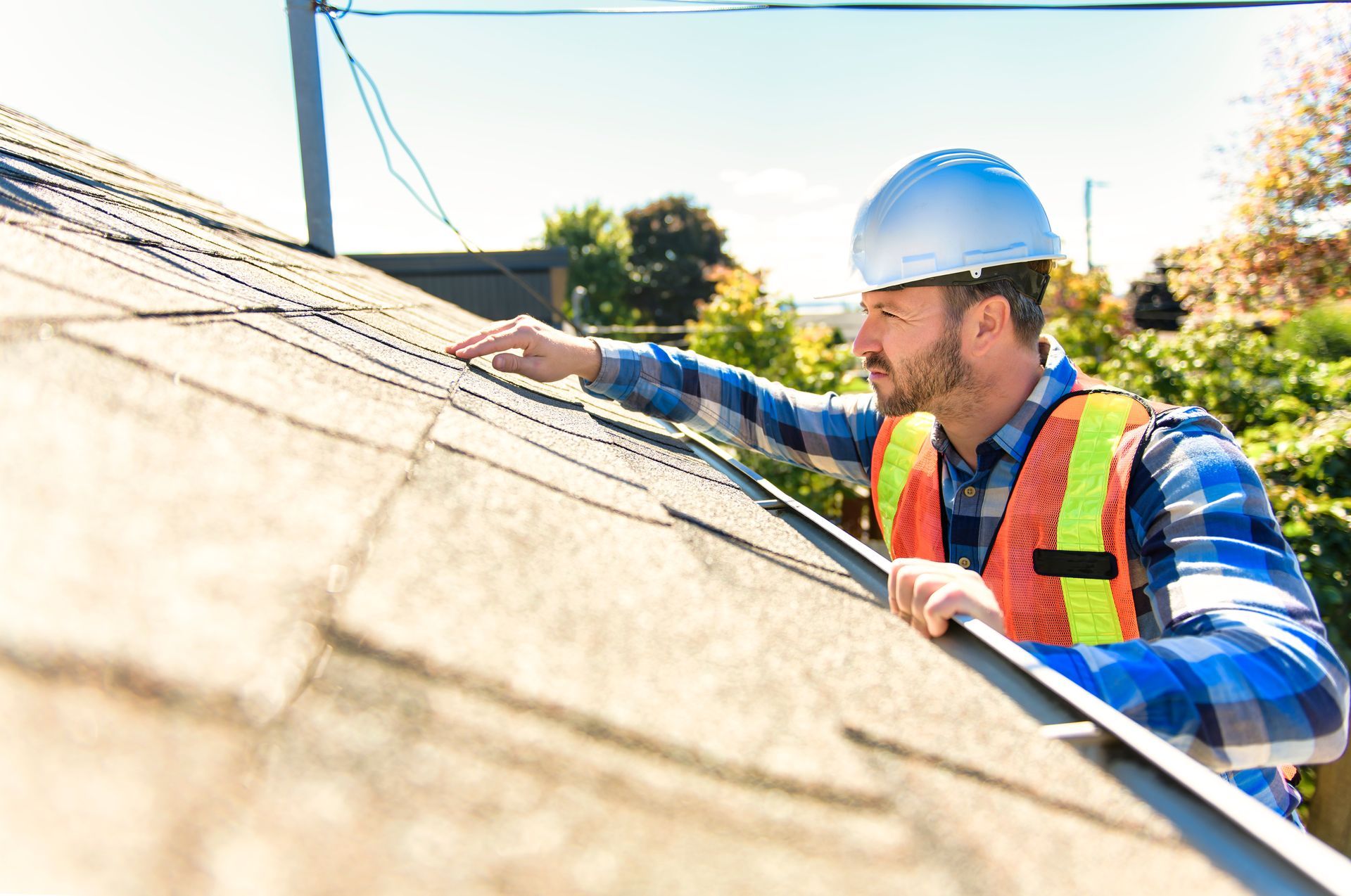 Roofer in hard hat and safety vest inspecting shingles on a rooftop on a sunny day.