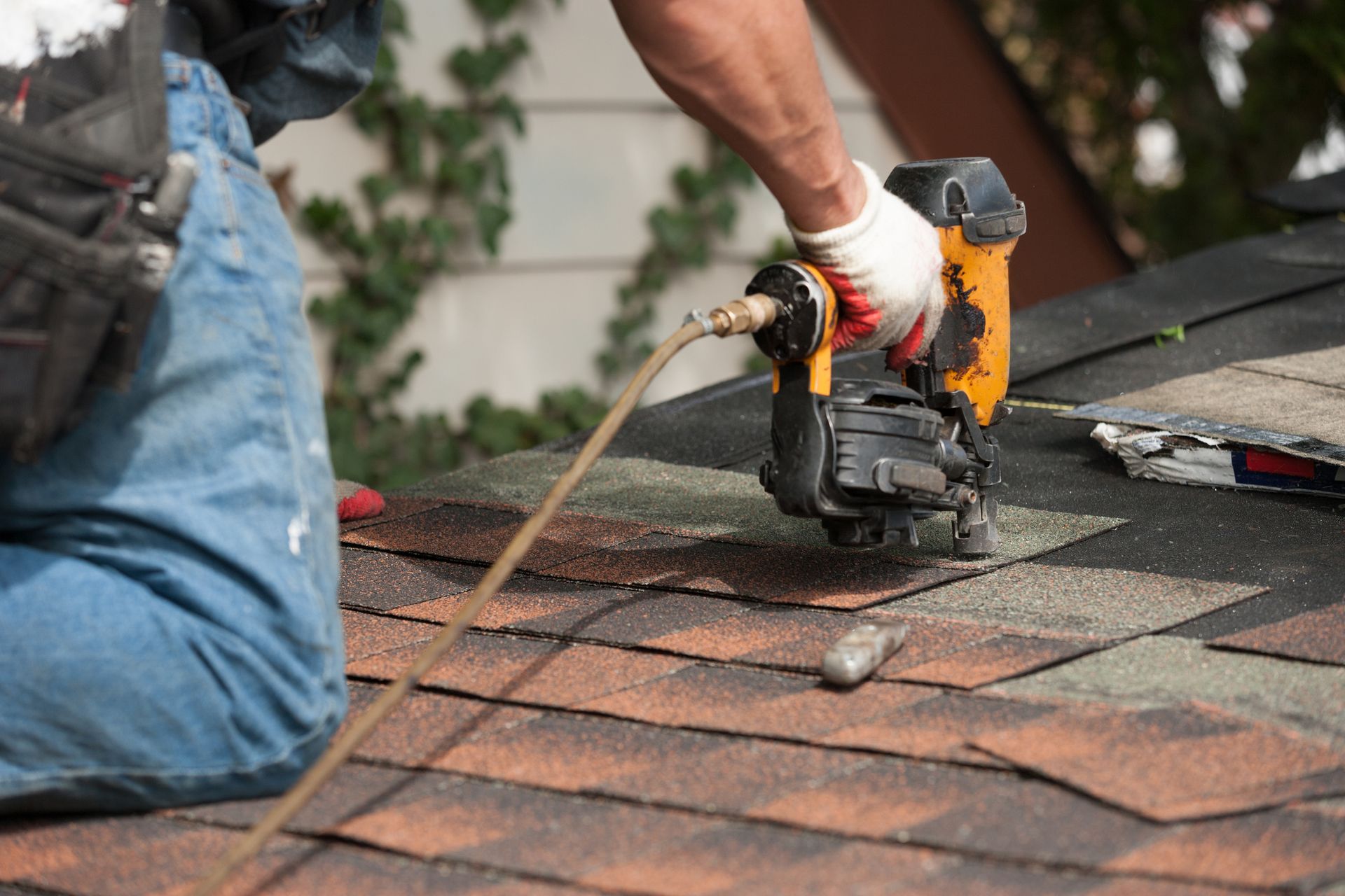 A roofing installation contractor is installing asphalt shingles on top of a roof with a nail gun.