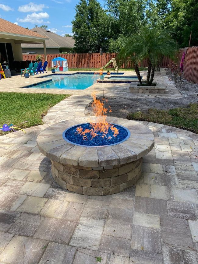 Backyard patio with a round stone fire pit, pool, trampoline, and trees in the background