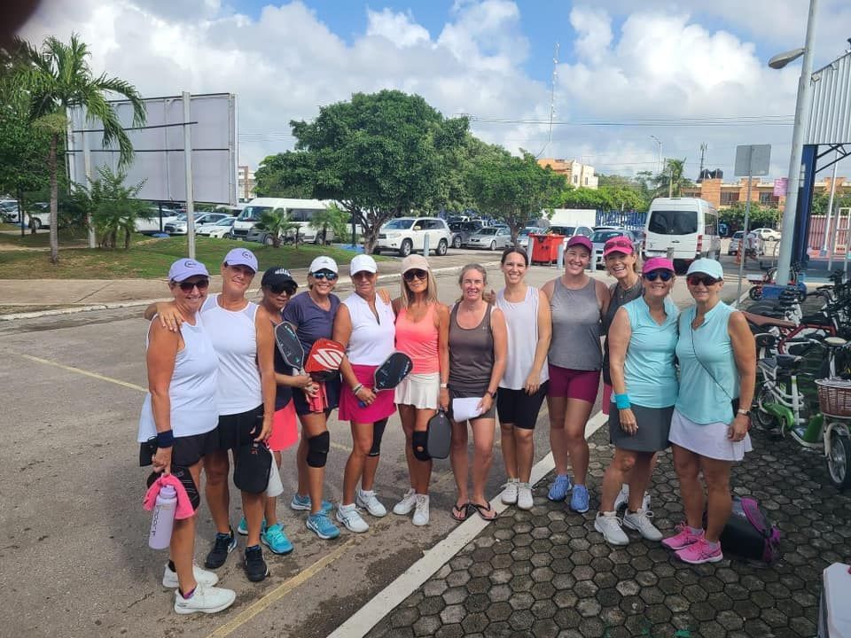 A group of women pickleball players are posing for a picture in a parking lot  at the Playa Del Carmen Pickleball Club in Playa Del Carmen, Mexico rackets.
