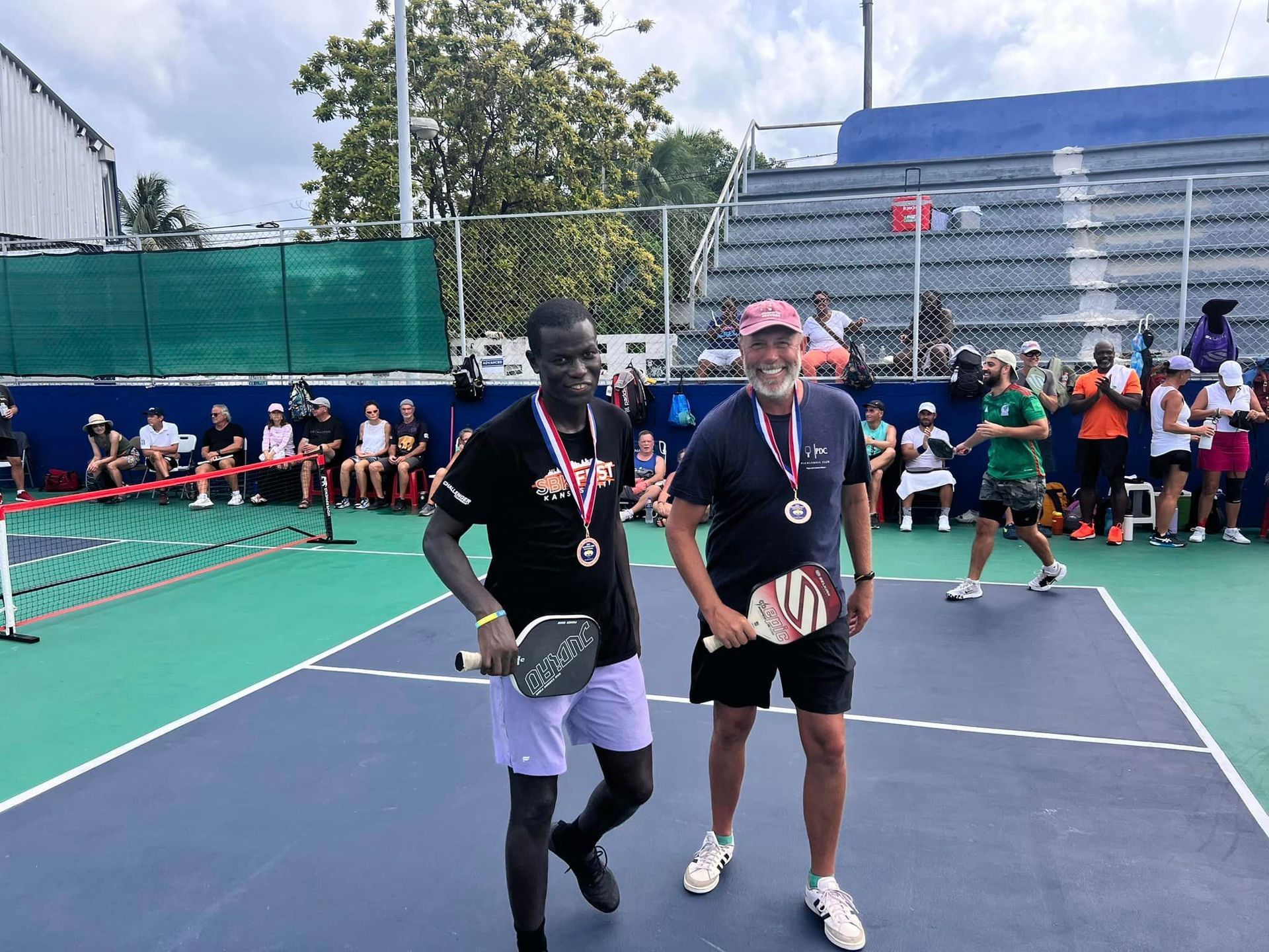 Two men are standing on a pickleball court with medals around their necks.  at the Playa Del Carmen Pickleball Club in Playa Del Carmen, Mexico rackets.