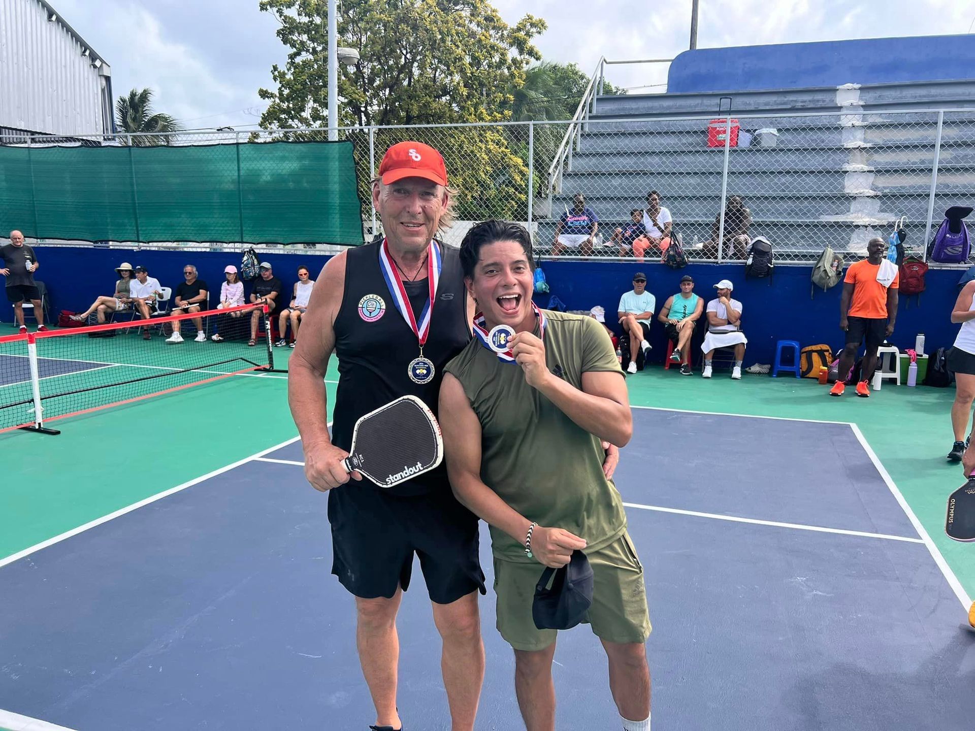 Two men are posing for a picture on a pickleball court  at the Playa Del Carmen Pickleball Club in Playa Del Carmen, Mexico rackets.