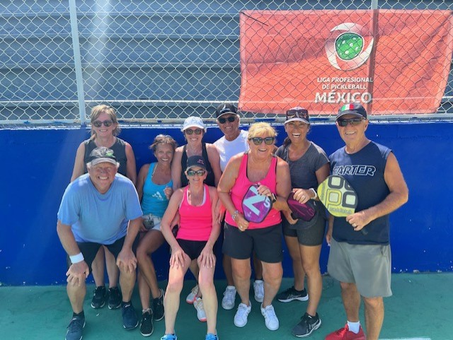 A group of PDC pickleball club members are posing for a picture on a tennis court in front of a banner that says mexico  at the Playa Del Carmen Pickleball Club in Playa Del Carmen, M