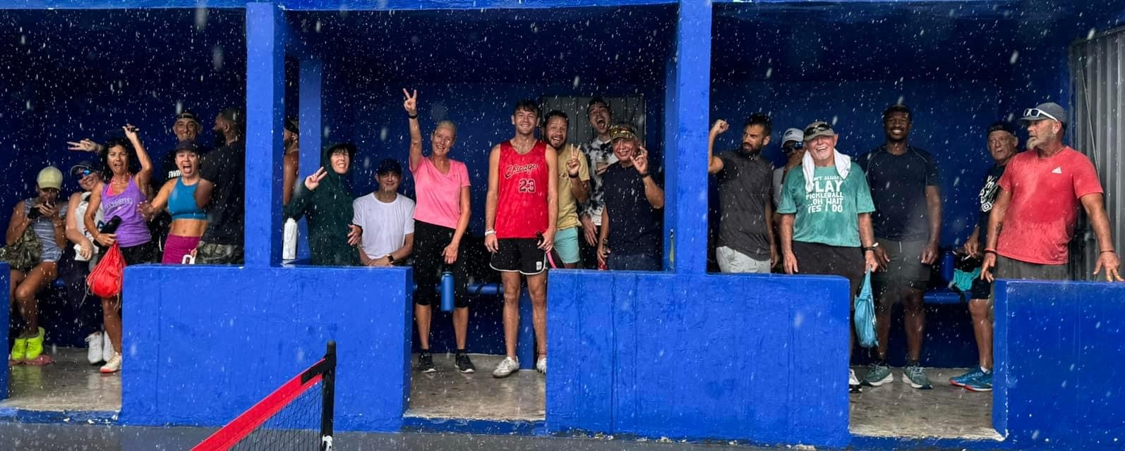 A group of people are standing in front of a blue wall  at the Playa Del Carmen Pickleball Club in Playa Del Carmen, Mexico rackets.
