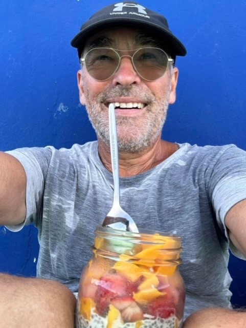 A man wearing sunglasses and a hat is holding a jar of fruit with a spoon in his mouth.