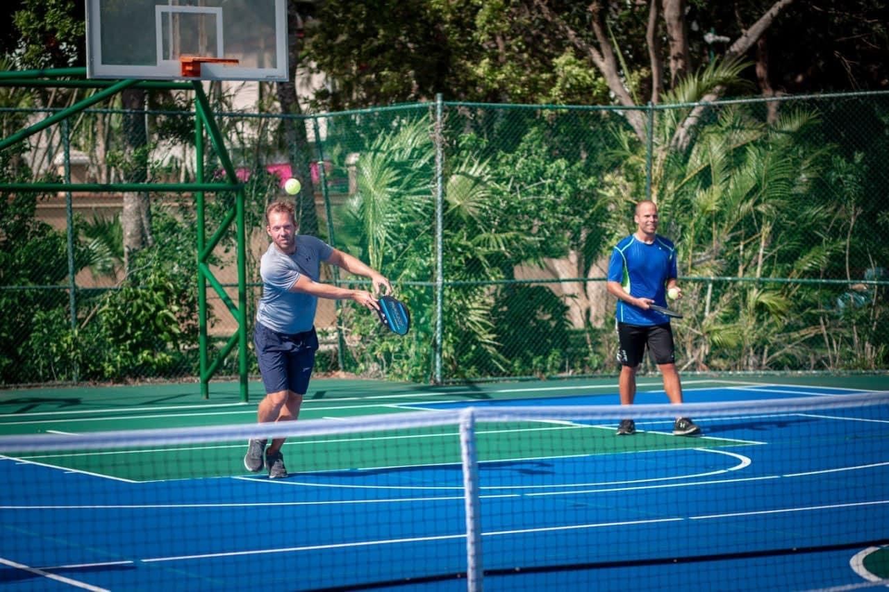 Two men are playing pickleball on a court with a basketball hoop in the background.