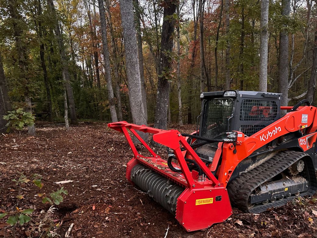 Orange Kubota track loader with mulching head in a wooded area, shredding ground cover.