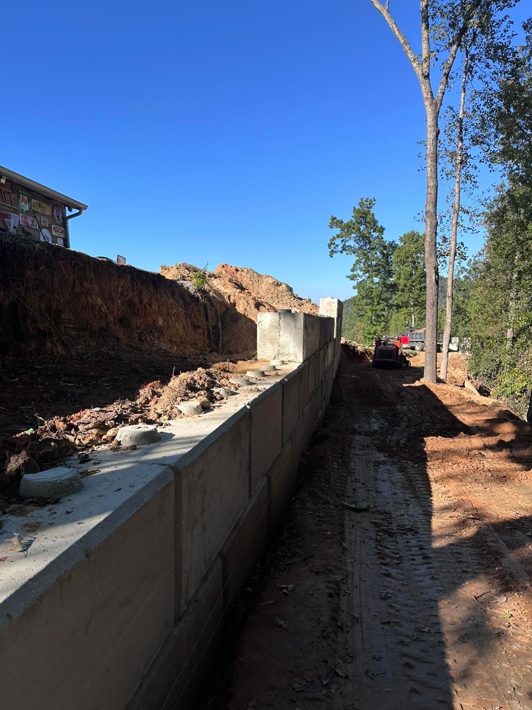 Concrete retaining wall construction on a hillside, with dirt, trees, and a clear blue sky.