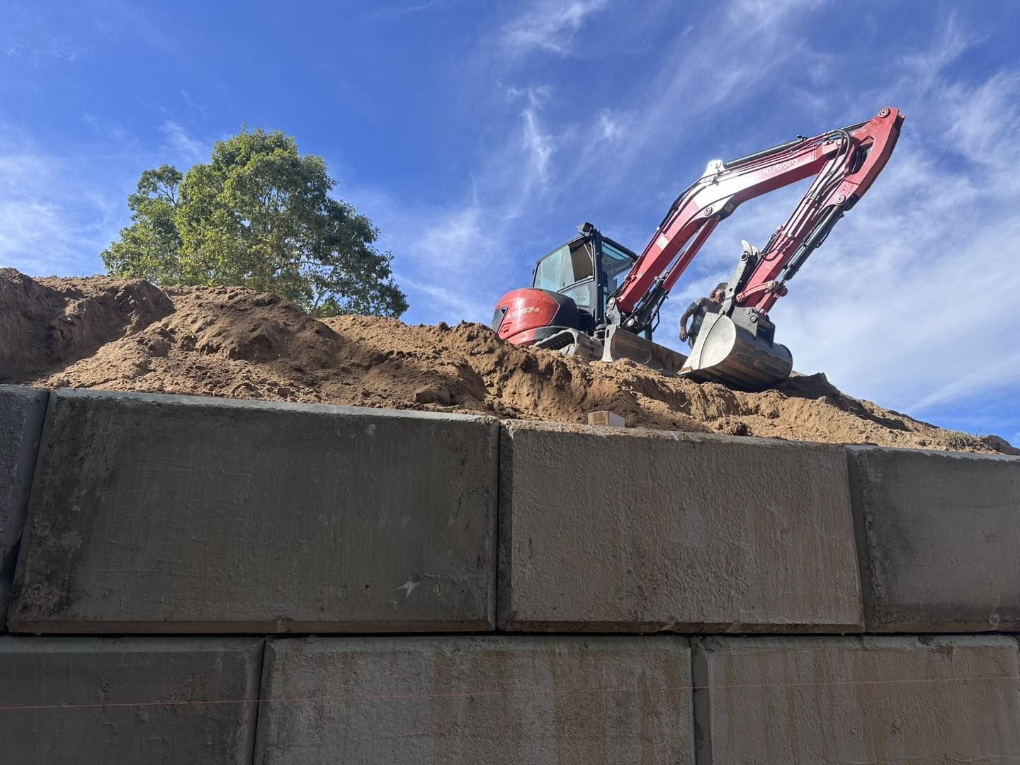 Excavator on a mound of dirt against a blue sky, working on a retaining wall made of concrete blocks.