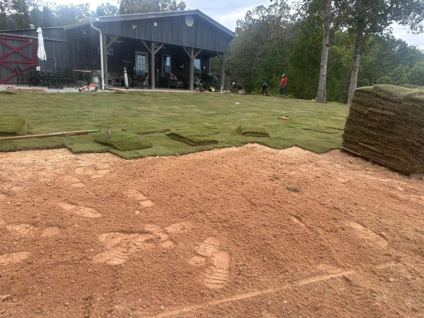 Laying sod in front of a wooden building. Green grass, sandy ground, and a stack of sod.