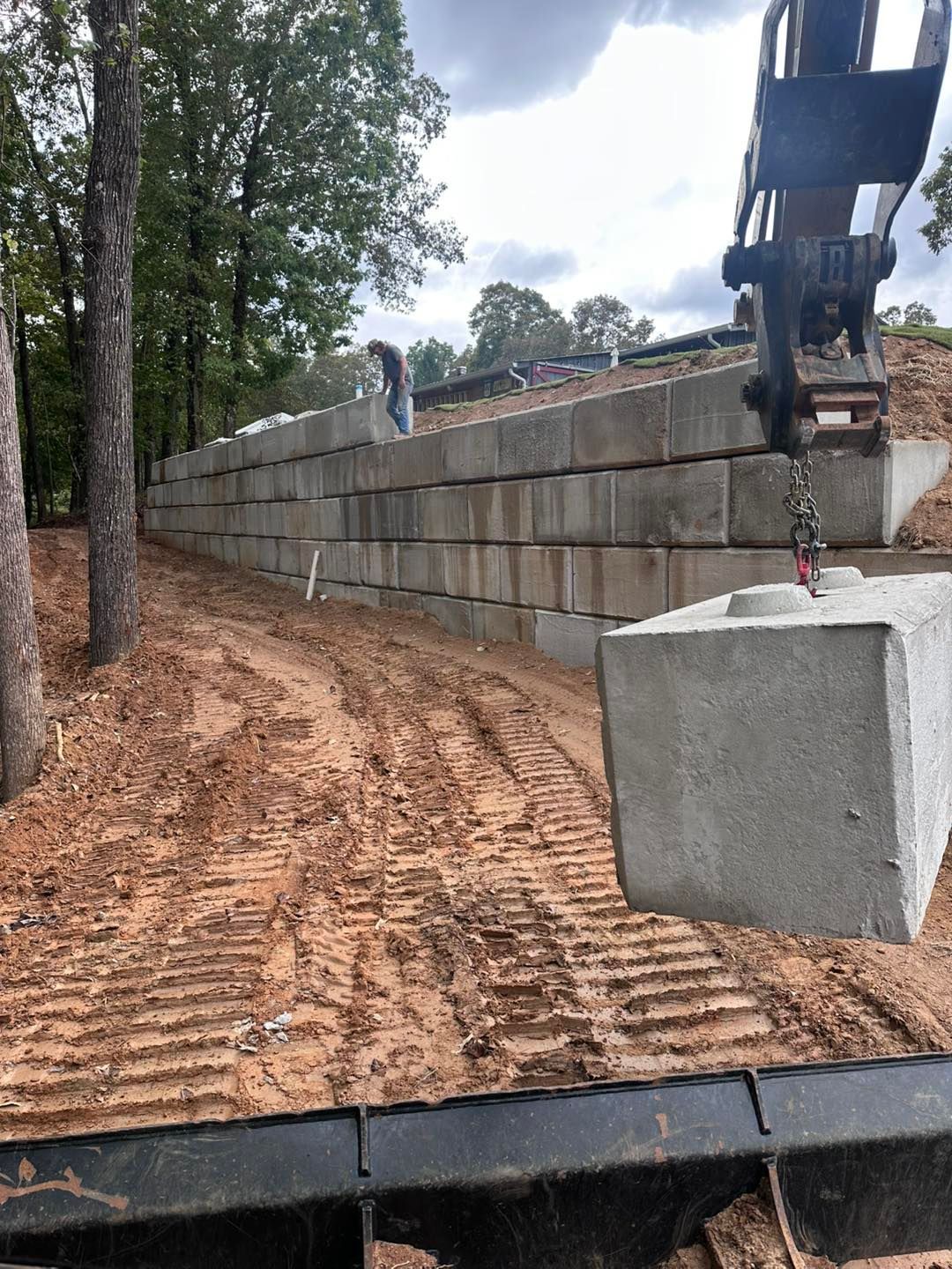 Excavator placing concrete block for a retaining wall on a dirt embankment near trees.