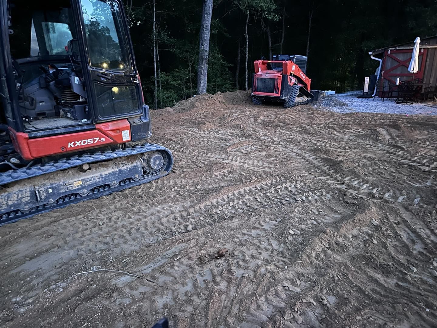 Excavator and skid steer on a dirt lot. Trees in background, with a small building.