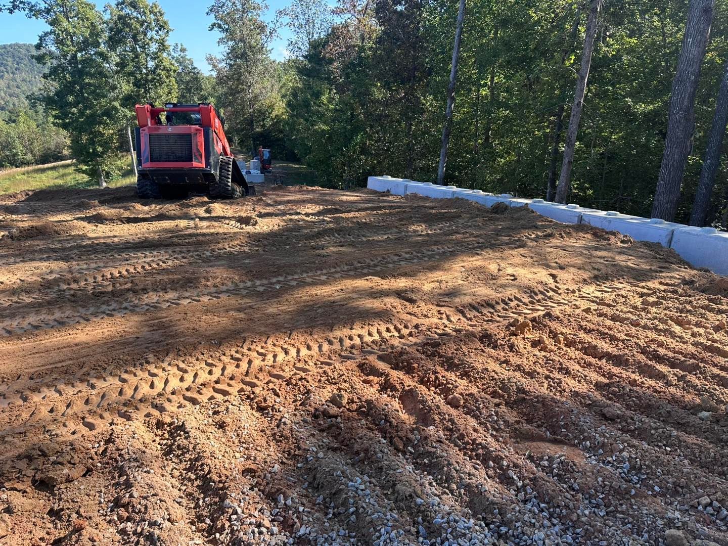 A red skid steer on a cleared plot of land with a retaining wall; forest in the background.