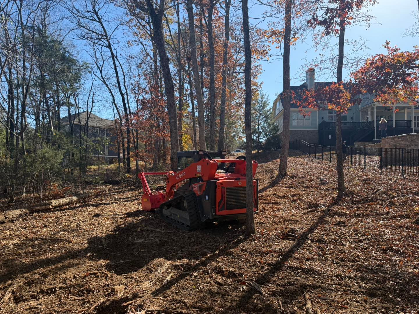 Orange skid steer on a leaf-covered hillside clearing leaves in a wooded area with houses visible.