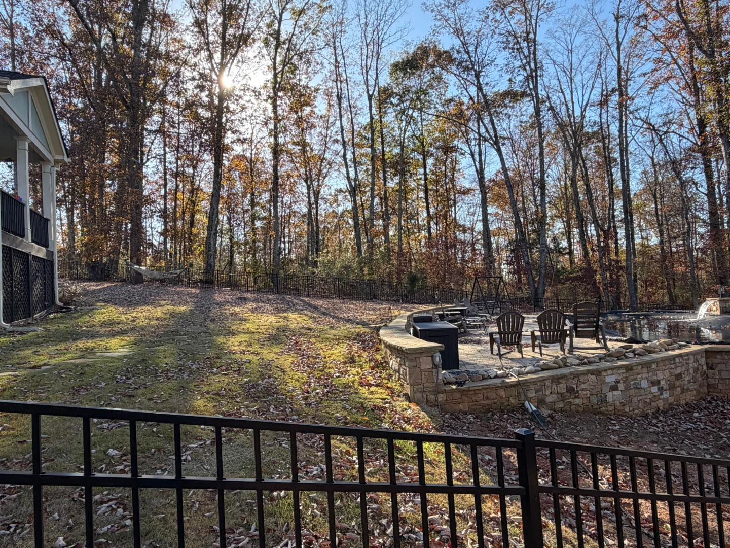 Backyard with trees, patio, retaining wall, and black fence on a sunny day.