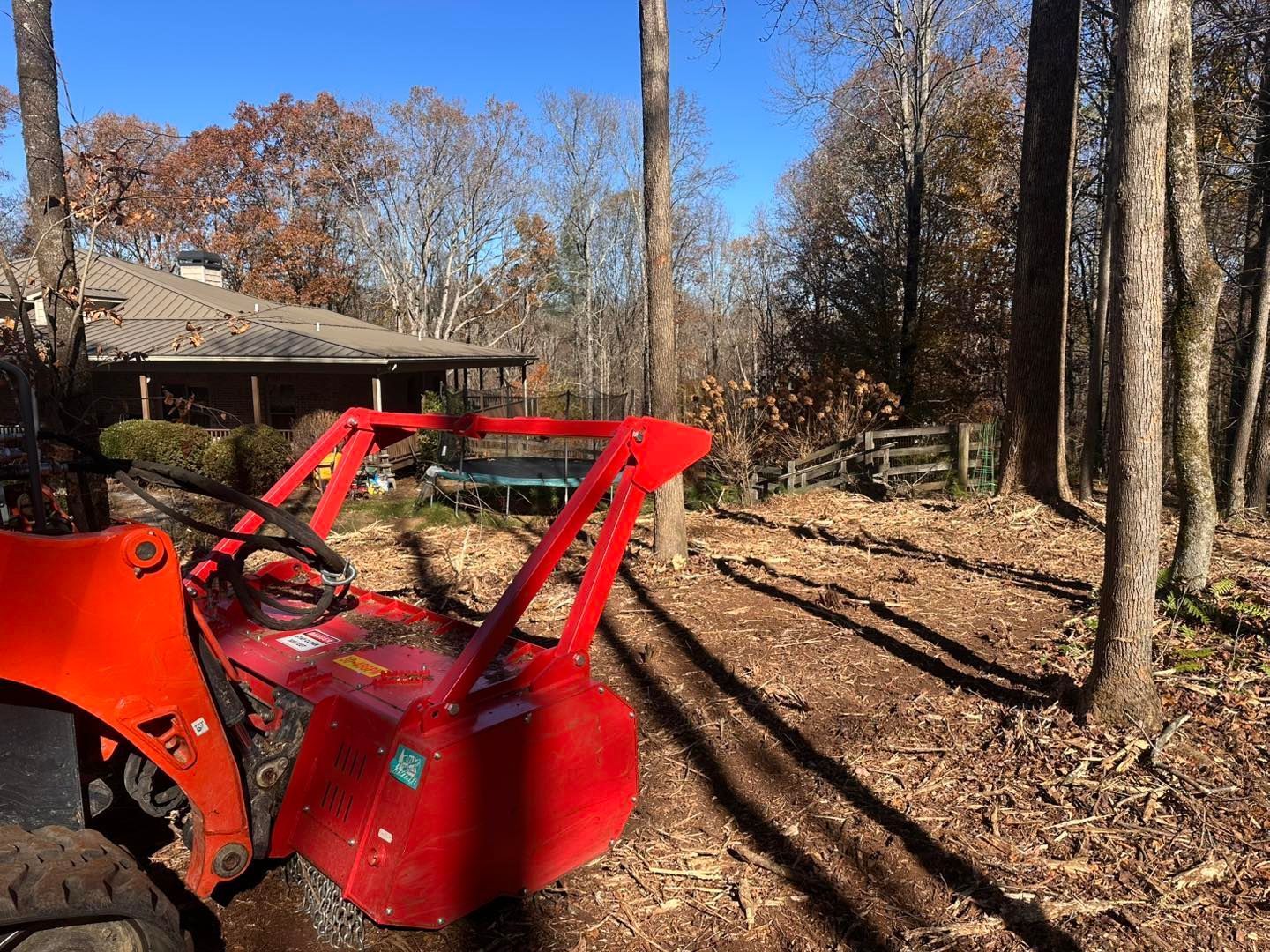 Red tractor with rotary tiller clearing a wooded area near a house.