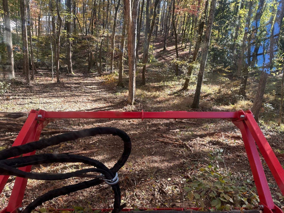 Red tractor front-end, foreground, facing a wooded area. Brown leaves cover the ground. Sunlight filters through the trees.